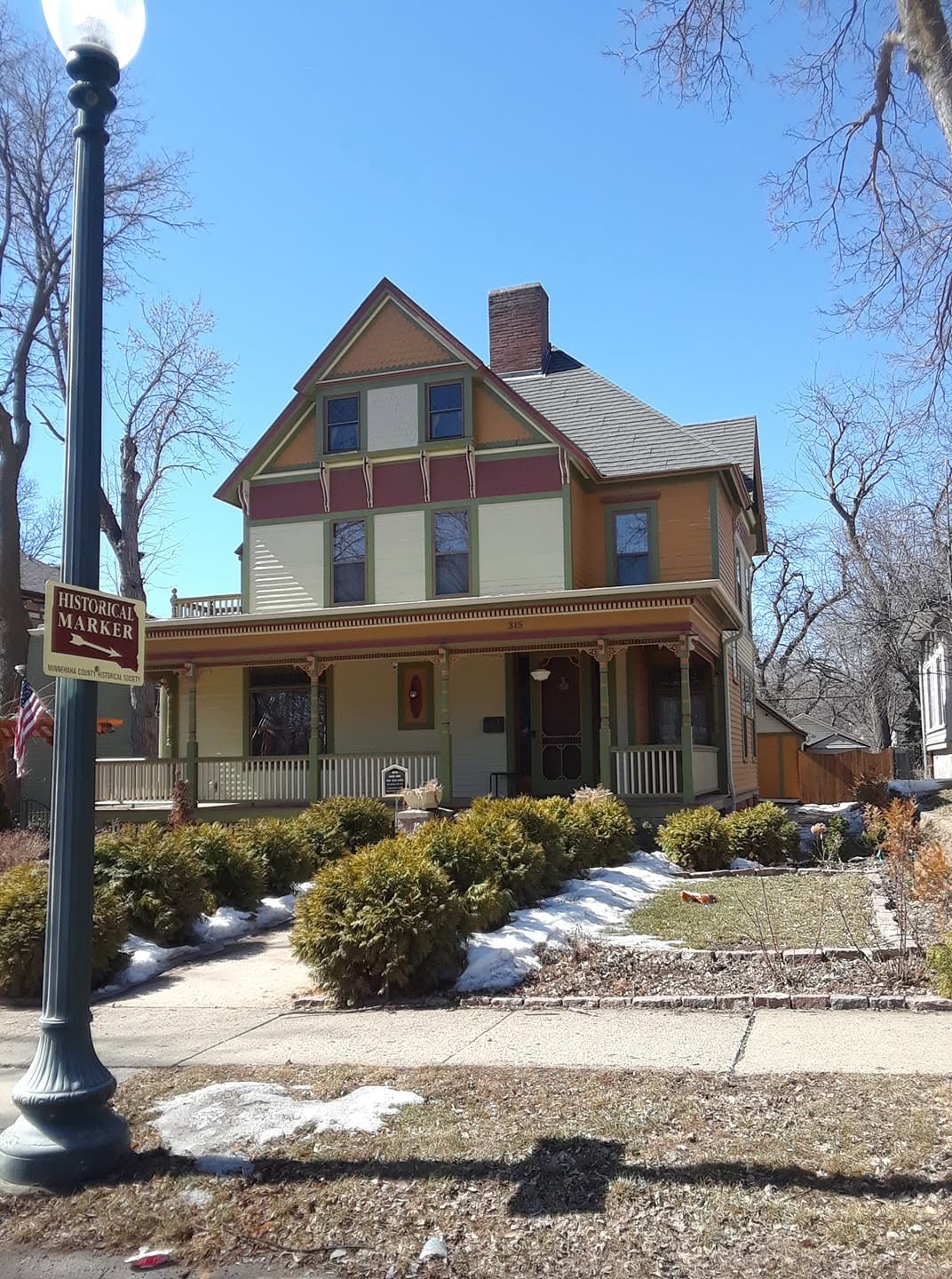 Two-and-a-half-story Victorian house freshly painted in mustard, sage, cream, and burgundy, ornate spindle porch, a historical-marker sign on the streetlight at the curb